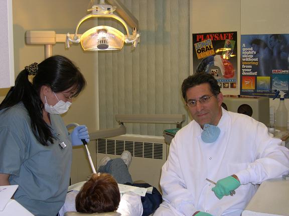 In the image, there are two individuals wearing medical scrubs and masks, with one person holding a tray containing a toothbrush and dental instruments. They appear to be in an indoor setting that resembles a dental or medical office, suggesting a dental hygiene or oral health education event.