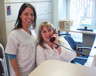 A woman wearing a white lab coat stands next to another woman who is on a phone at an office desk.