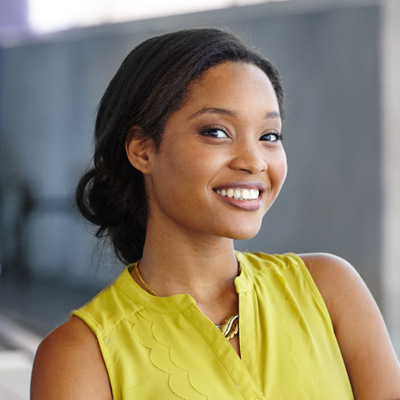 A young woman with a bright smile, wearing a yellow blouse, poses for a portrait against a blurred background.