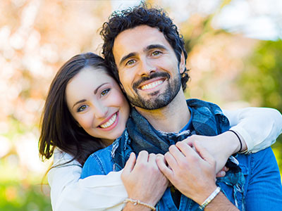 A man and woman are embracing each other, smiling, with the man wearing a bandana around his neck and both dressed casually, against a blurred background that suggests an outdoor setting.