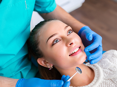 In the image, there s a female patient sitting in a dental chair with a smile on her face, receiving dental care from a dentist who is adjusting her teeth with a tool.