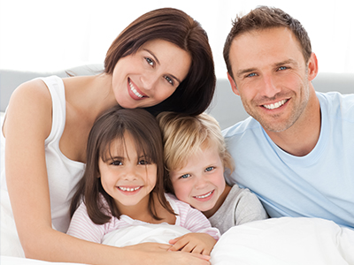 The image shows a family of four people, including two adults and two children, all smiling and posing together on a bed.