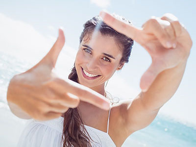 The image features a smiling woman with long hair, holding her hand up to her face as if taking a selfie, against a bright background with clear skies.